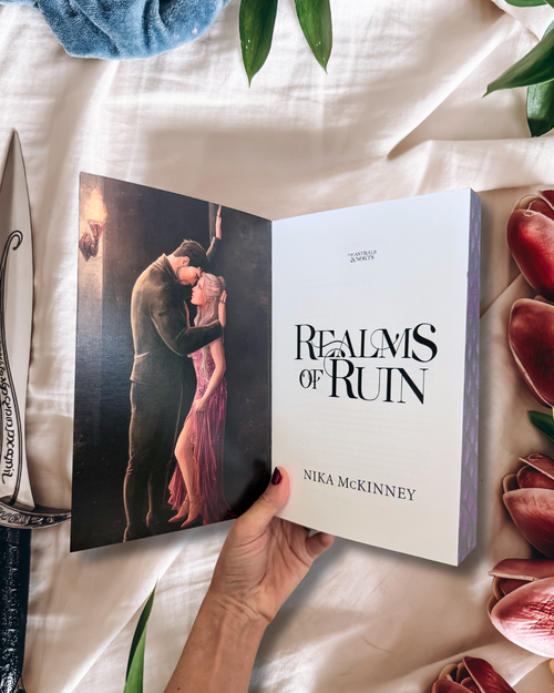 A hand holds an open book titled "Realms of Ruin" by Nika McKinney, showing a romantic illustration on the left page and the white title page on the right. The book is photographed on draped white fabric with decorative leaves, flowers, and a blue cloth around it.
