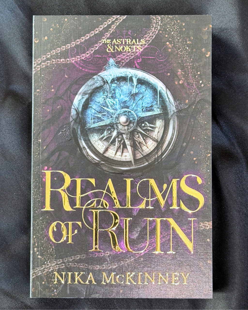 The book cover of 'Realms of Ruin' by Nika McKinney, featuring a detailed fantasy illustration of a compass with a blue sky and purple clouds, displayed on a dark fabric background.