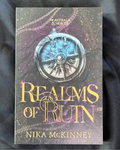 The book cover of 'Realms of Ruin' by Nika McKinney, featuring a detailed fantasy illustration of a compass with a blue sky and purple clouds, displayed on a dark fabric background.